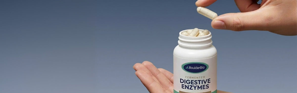 Hand removing a capsule from a bottle of BoulderBio Therapeutic Strength Digestive Enzymes with a blue gradient background.